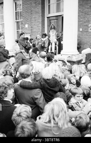 Empfang des Heiligen Nikolaus in Heemskerk St. Nikolaus Sinterklaas, Whizgle News, Dutch Desk, Niederlande, 1950 - 2000 am 22-11-1969. Diese Themen sind in der Abbildung dargestellt. Die Szene ist voller Aktivität, die sich um eine Gruppe von Personen dreht, die sich vor einem großen Eingang versammelt haben, der mit hohen weißen Säulen geschmückt ist. Das Gebäude hat eine Ziegelfassade, die ein Gefühl von Tradition und Geschichte vermittelt. An vorderster Front ist eine große Menschenmenge dicht gepackt, ihre Ausdrucksformen sind eine Mischung aus Spannung und Vorfreude. Die Menge ist vielfältig, mit Menschen unterschiedlichen Alters, einschließlich Kindern mit Tous Stockfoto