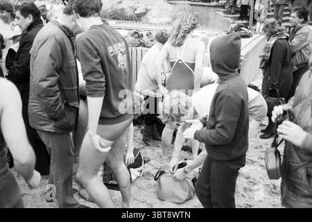 Silvester Tauchgang Zandvoort Neujahr Zandvoort, Whizgle News, Dutch Desk, Niederlande, 1950 - 2000 am 01-01-1984. Das Bild enthält diese Themen. Die Szene fängt einen lebendigen Moment am Strand ein, gefüllt mit einer Mischung aus Aktivität und Vorfreude. Die Menschen sind eng zusammengekommen und schaffen ein Gefühl von Gemeinschaft und Kameradschaft. Im Vordergrund bereiten sich Individuen in Badeanzügen auf das Schwimmen vor, deren Körper teilweise von Handtüchern und Taschen verdeckt sind, die auf dem Sandboden verstreut sind. Die Badeanzüge sind in verschiedenen Stilen erhältlich, einige sind einteilig, andere sind sportlicher. Eine Person, die einen eng anliegenden Anzug trägt Stockfoto