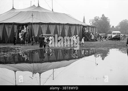 Überschwemmung um Haarlem Flood, Whizgle News, Dutch Desk, Niederlande, 1950 - 2000 am 07-08-1968. Die Abbildung zeigt diese Themen. Die Szene zeigt eine lebendige Zirkusumgebung vor einem Hintergrund aus grauem Himmel und gedämpften Tönen. Die zentrale Struktur ist ein großes, klassisches Zirkuszelt mit einem charakteristischen, konischen Gipfel, der mit farbenfrohen Stoffen in wechselnden blau-roten Paneelen verziert ist. Das Zelt steht auf leicht unebenem Boden, umgeben von Pfützen, die seine leuchtenden Farben widerspiegeln. Es werden Gruppen von Menschen beobachtet, die sich bewegen, einige steigen auf und ab, während andere wandern Stockfoto
