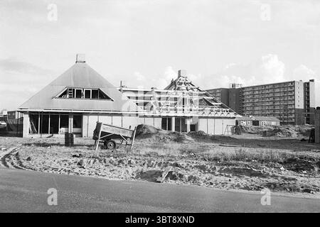 Villen im Zentrum von Heemstede Construction sites, Whizgle News, Dutch Desk, Niederlande, 1950 - 2000 am 25-01-1972. Das Bild enthält diese Themen. In der Szene werden zwei Strukturen hervorgehoben dargestellt, die unterschiedliche Architekturstile zeigen. Auf der linken Seite befindet sich ein teilweise erbautes Gebäude mit einem steilen, dreieckigen Dach und großen Fenstern, die einen Einblick in die Innenkonstruktion ermöglichen. Die Mauern sind aus Ziegeln gefertigt und ein Fundament befindet sich noch in der Fertigstellung. Auf der rechten Seite erhebt sich ein moderneres Hochhaus mit einer einheitlichen Fassade mit zahlreichen Windowänden Stockfoto