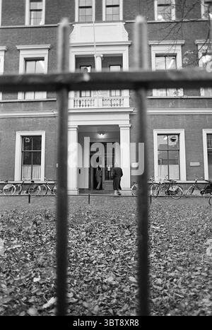 Regional Labor Office Haarlem Nederland., Whizgle News, Dutch Desk, Niederlande, 1950 - 2000. Diese Themen sind in der Abbildung dargestellt. Die Szene zeigt ein markantes Gebäude mit klassischem Architekturstil mit roten Ziegelwänden und großen Fenstern, die von weißen Zierleisten eingerahmt sind. Der Eingang ist durch eine breite Reihe von Stufen gekennzeichnet, die zu einer großen Tür führen, die durch dekorative Elemente oberhalb und flankiert wird. Im Vordergrund trennt ein schwarzer schmiedeeiserner Zaun den Betrachter vom Eingang und verleiht der Komposition ein Gefühl von Tiefe. Die Gitter des Zauns erzeugen ein Muster, das Stockfoto