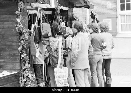 Ostermassen in Zandvoort und Verkehrsstau Verspronckweg geschäftig, Whizgle News, Dutch Desk, Niederlande, 1950 - 2000 am 16-04-1979. Diese Themen werden in der Abbildung angezeigt. Die Szene ist voll von Aktivitäten an einem Marktstand, gekennzeichnet durch eine Holzstruktur mit verschiedenen Gegenständen zum Verkauf. Es sind mehrere Personen versammelt, die die Angebote untersuchen. Die Figuren sind vielfältig im Aussehen, mit verschiedenen Frisuren und Kleidungsstilen, die eine Mischung aus lässiger und leicht Retro-Mode widerspiegeln. Die Menschen stehen in unmittelbarer Nähe und schaffen ein Gefühl der gemeinsamen Interaktion. Einige sind lea Stockfoto