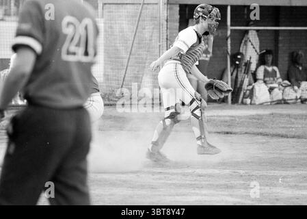 Softball Bloemendaal - DSC, Whizgle News, Dutch Desk, Niederlande, 1950 - 2000 am 05-05-1990. Dies sind die Themen im Bild. In der Szene findet ein lebhaftes Softballspiel auf einem gut gepflegten Feld statt, das durch sattes grünes Gras und ein staubiges Infield gekennzeichnet ist. Die Atmosphäre ist lebhaft, voller Energie des Wettbewerbs. Eine junge weibliche Fängerin, die im Vordergrund steht, trägt eine traditionelle Uniform mit Streifen. Ihre Schutzausrüstung glitzert im Sonnenlicht und reflektiert verschiedene Schwarz- und Grautöne. Sie steht bereit, leicht gehockt, konzentriert Stockfoto
