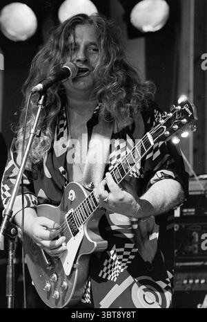 Red Cross Pop Konzert Grote Markt Haarlem Grote Markt The Netherlands, Whizgle News, Dutch Desk, Niederlande, 1950 - 2000 am 28.05.1993. Das Bild enthält diese Themen. In einer lebendigen Performance-Umgebung steht ein Musiker im Mittelpunkt der Bühne, der vom Rhythmus der Musik fasziniert ist. Sein langes, welliges Haar fällt über seine Schultern und reflektiert das Licht mit einem natürlichen Glanz. Er hält eine E-Gitarre mit einem satten, leuchtenden Finish, die warme Gold- und Brauntöne vereint. Die Kleidung des Musikers ist auffällig und zeichnet sich durch auffällige Muster in Schwarz, weiß und tiefen Farben aus, die ein eklektisches s widerspiegeln Stockfoto