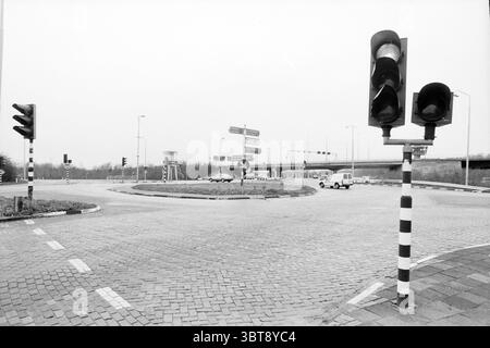 Überquerung von Oude Schipholweg 'Kreisverkehr' Straßen Straßenbau Straßenbau Strassenbau Schipholweg, Whizgle News, Dutch Desk, Niederlande, 1950 - 2000 am 29-12-1986. Die Abbildung zeigt diese Themen. Die Szene zeigt einen Kreisverkehr in gedämpftem, monochromatischem Ton, der stark auf Grauschattierungen ausgerichtet ist. Im Vordergrund stehen verschiedene Ampeln, wobei die grünen Ampeln auf der linken Seite sanft vor der bewölkten Kulisse leuchten. Die Ampeln sind auf gestreiften Stangen montiert, um die Sichtbarkeit in den umliegenden dunklen Farben zu verbessern. Um den Kreisverkehr herum befinden sich mehrere Straßenschilder, die auf den Kreisverkehr hinweisen Stockfoto
