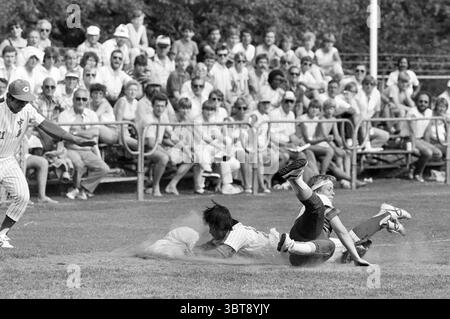 Niederlande - Taiwan Softball, Whizgle News, Dutch Desk, Niederlande, 1950 - 2000 am 10-07-1983. Das Bild enthält diese Themen. Die Szene fängt einen lebendigen Moment während eines Baseballspiels an einem hellen Tag ein. Im Vordergrund gleiten zwei Spieler in eine Basis, ihre Körper in Bewegung und erzeugen ein Gefühl von Spannung und Spannung. Ein Spieler trägt eine weiß gestreifte Uniform mit fokussiertem Ausdruck, während der andere, in dunkler Kleidung gekleidet, ihn wahrscheinlich auszuspielen versucht. Beide Spieler sind von einer Staubwolke umgeben, die vom Spielfeld aufsteigt und die Action unterstreicht. Hintergrunddetails zeigen einen Pel Stockfoto
