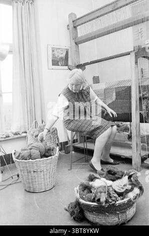 Raum für Kreativität mit Drehrad Webcollagen Mann Malerei und Frau, die auf großen Wandteppichen oder Teppichen arbeitet.., Whizgle News, Dutch Desk, Niederlande, 1950 - 2000. Diese Themen sind in der Abbildung dargestellt. Die Szene präsentiert ein gemütliches Interieur mit einer warmen, kreativen Atmosphäre. Eine Frau sitzt auf einem Stuhl neben einem großen Webstuhl, die sich aktiv in ihrem Handwerk engagiert. Sie hat helles Haar und trägt ein gemustertes Kleid mit weißem Oberteil, das ihrem Aussehen einen Hauch von Tradition verleiht. Der Webstuhl selbst ist deutlich sichtbar platziert und zeigt ein teilweise gewebtes Textil, das auf komplizierte Designs und Texte hinweist Stockfoto