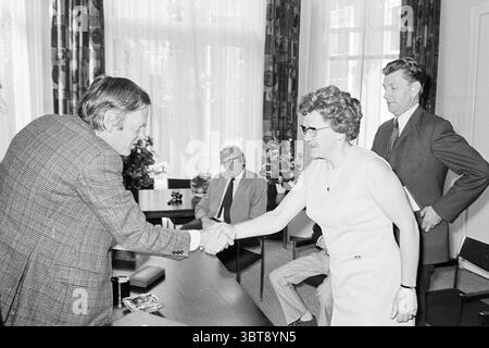 Drei Schullehrer zu ehren. Ehren, Whizgle News, Dutch Desk, Niederlande, 1950 - 2000 am 23-06-1972. Das Bild enthält diese Themen. Die Szene verfügt über eine formelle Inneneinrichtung, die durch einen großen, gut beleuchteten Raum gekennzeichnet ist. Sonnenlicht filtert durch große Fenster, wirft weiche Schatten auf den Boden und verbessert die Helligkeit insgesamt. Im Vordergrund stehen sich zwei Personen in einem engagierten Handschlag zueinander gegenüber. Eine Person, die in einem strukturierten Karomuster gekleidet ist, scheint mittleren Alters zu sein und eine Brille trägt. Die andere ist eine Frau in einem ärmellosen, hellen Kleid mit einer selbstbewussten Haltung Stockfoto
