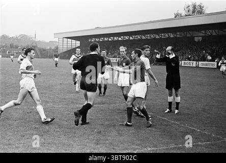 VSV - SHS Football, Whizgle News, Dutch Desk, Niederlande, 1950 - 2000 am 24-09-1961. Dies sind die Themen im Bild. Die Szene fängt einen dynamischen Moment auf einem Sportplatz ein, wahrscheinlich während eines Fußballspiels. Die Spieler sind auf dem grasbewachsenen Spielfeld verstreut, wobei mehrere in weißen Trikots und andere in dunklen Trikots gekleidet sind, was die beiden konkurrierenden Teams angibt. Die Spieler in weiß scheinen sich in einem Handschlag mit denen in dunklerer Kleidung zu befinden, was auf einen Moment sportlicher Geschicklichkeit oder vielleicht auf einen Abschluss des Spiels hindeutet. Die Einstellung verfügt über einen großen überdachten Ständer im Hintergrund, gefüllt Stockfoto