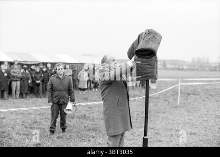 Enthüllung des Namensschilds Hindernislauf für Hunde bei MSD Dogs Reveads, Whizgle News, Dutch Desk, Niederlande, 1950 - 2000 am 21.12.1988. Die Abbildung zeigt diese Themen. Ein Mann in einem langen Mantel steht im Vordergrund, der eine schwere, dunkle Jacke hoch über dem Kopf hebt. Sein Ausdruck ist Enthusiasmus oder Entschlossenheit und deutet auf einen Moment der Bedeutung oder des Feierns hin. Die Jacke bildet einen Kontrast zu den gedämpften Tönen des bewölkten Himmels, der ein weiches, gleichmäßiges Licht über die Szene wirft und ein Gefühl von Ruhe und doch ernster Atmosphäre schafft. Im Hintergrund versammeln sich eine Gruppe von Menschen Stockfoto
