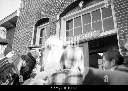 Weihnachtsmann vor dem Bahnhof Bloemendaal Weihnachten Weihnachtsdekoration Weihnachtsdekoration Weihnachtsdekoration, Whizgle News, Dutch Desk, Niederlande, 1950 - 2000 am 27-12-1962. Diese Themen sind in der Abbildung dargestellt. Die Szene fängt ein lebhaftes Treffen am Eingang eines Gebäudes ein, das durch seine rote Backsteinfassade gekennzeichnet ist. Die Architektur verfügt über große, bogenförmige Fenster, die natürliches Licht hereinlassen und ein ruhiges Leuchten auf die Gesichter der umliegenden Personen werfen. Eine festliche Atmosphäre ist offensichtlich, verstärkt durch die Anwesenheit einer Figur, die als Weihnachtsmann verkleidet ist, der mit seinem langen weißen Bären große Aufmerksamkeit auf sich zieht Stockfoto