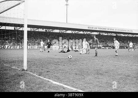 Telstar - Ajax Football Telstar, Whizgle News, Dutch Desk, Niederlande, 1950 - 2000 auf 30-04-1972. Diese Themen sind in der Abbildung dargestellt. Die Szene fängt einen intensiven Moment eines Fußballspiels in einem Stadion voller Zuschauer ein. Das Feld ist ein sattes, lebendiges Grün, das sich von den gedämpften Tönen des bewölkten Himmels abhebt. Im Vordergrund taucht ein Torhüter in einer auffälligen roten Uniform nach links, die Arme ausgestreckt, während ein Ball auf das Tor zusteuert. Sein Ausdruck vermittelt Entschlossenheit und Dringlichkeit. Direkt hinter ihm zeigen Verteidiger in weißen Trikots eine Mischung aus Vorfreude und Concept Stockfoto
