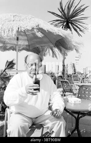 Simon van Collem with a Drink Personen t.v., Whizgle News, Dutch Desk, Niederlande, 1950 - 2000 am 18-06-1983. Die Abbildung zeigt diese Themen. Die Szene fängt eine sonnendurchflutete Umgebung im Freien ein, wahrscheinlich am Strand oder an einer belebten Promenade. Im Vordergrund sitzt ein Mann mittleren Alters mit rundem Gesicht und Brille an einem kleinen runden Tisch. Er trägt ein helles Sweatshirt, das eine entspannte Atmosphäre vermittelt. Der Mann wiegt ein hohes Glas, gefüllt mit einem dunklen Getränk, geschmückt mit einer Scheibe Zitrone und möglicherweise einem Strohhalm, was ein erfrischendes Getränk an einem warmen Tag andeutet. Er umgibt lebendige Elemente des Sommers. A Large Stockfoto