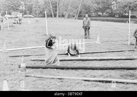 Festlichkeiten IJmuiden Beverwijk Haarlem Queen's Day, Whizgle News, Dutch Desk, Niederlande, 1950 - 2000 auf 30-04-1974. Dies sind die Elemente im Bild. In einer grasbewachsenen Umgebung im Freien wird eine Gruppe von Kindern spielerisch aktiv. Zwei Kinder stehen im Vordergrund: Eines springt energisch über einen Baumstamm, während sich ein anderes hinunterhockt und sich scheinbar auf das Klettern oder Navigieren konzentriert. Beide Kinder tragen legere Kleidung, die für Outdoor-Spiele geeignet ist, wobei eines ein dunkles Oberteil und das andere ein leichteres Outfit trägt. Um sie herum liegen mehrere Holzpfähle Stockfoto
