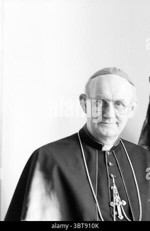 Bischof Bomers Roman Catholic Church, Whizgle News, Dutch Desk, Niederlande, 1950 - 2000 am 23-02-1989. Diese Themen werden in der Abbildung angezeigt. In der Szene ist ein Mann in traditioneller Kleruskleidung zu sehen. Seine Kleidung ist meist dunkel, mit einem dezenten Glanz, der auf einen hochwertigen Stoff hinweist. Ein langer, fließender umhang oder Keuschel reicht bis zu den Schultern, mit einem markanten Kragen, der das formelle Erscheinungsbild unterstreicht. Er trägt eine Brille, die sein Gesicht umrahmt und ein Gefühl von Wärme und Zugänglichkeit vermittelt. Sein Ausdruck ist freundlich, mit einem sanften Lächeln, das Freundlichkeit und Offenheit vermittelt. Stockfoto