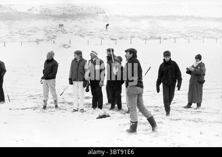 Golf am Noordwijk Beach - Zandvoort Golf Golf Golf Noordwijk, Whizgle News, Dutch Desk, Niederlande, 1950 - 2000 am 22-02-1986. Diese Themen sind in der Abbildung dargestellt. Die Szene zeigt einen Winterausflug an einem verschneiten Strand, an dem sich eine Gruppe von Individuen versammelt. Jede Person ist warm gekleidet und trägt schwere Mäntel, Hüte und Handschuhe, was auf das kalte Wetter hinweist. Ihre Gesichter zeigen eine Mischung aus Konzentration und Vergnügen, was auf eine gemeinsame Aktivität oder ein gemeinsames Ereignis hindeutet. Im Vordergrund ist ein Mann aktiv beschäftigt, mit einem langen Gegenstand in der Hand, als würde er sich darauf vorbereiten, etwas zu werfen oder zu werfen. Sein stabiler Fuß Stockfoto