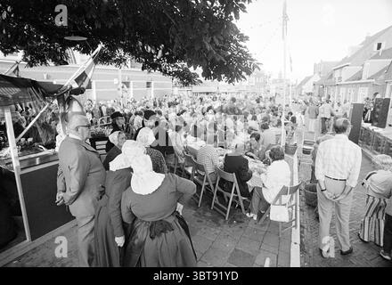 Fischmahlzeit unter freiem Himmel in Zandvoort Essen und Lokale Tourismus Touristen, Whizgle News, Dutch Desk, Niederlande, 1950 - 2000 am 18-07-1974. Das Bild enthält diese Themen. Die Szene ist voller Aktivität, bevölkert von einer großen Menschenmenge, die einen lebhaften Treffpunkt im Freien füllt. Die Menschen tragen eine Mischung aus traditioneller und zeitgenössischer Kleidung, einige tragen besondere historische oder kulturelle Kleidung mit Tüchern und Hauben. Die Farbpalette ist eine Mischung aus gedämpften Tönen; Pastelltöne sind neben tieferen Farben zu sehen, was eine einladende, aber leicht nostalgische Atmosphäre schafft. In der Zukunft Stockfoto