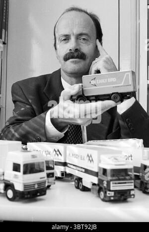 Herr Vallenduuk Transportanwalt mit Abholung von Miniatur-Lkw, Whizgle News, Dutch Desk, Niederlande, 1950 - 2000 am 18. 10. 1993. Die Abbildung zeigt diese Themen. Die Szene zeigt einen Mann mit einem auffälligen Schnurrbart, der in einem dunklen Anzug mit Krawatte gekleidet ist und selbstbewusst in einer Büroumgebung sitzt. Er steht an einem Tisch, der von mehreren Miniatur-Lkw überlagert ist, die alle das Design widerspiegeln, das man häufig im gewerblichen Verkehr sieht. Der Mann hält ein bestimmtes Modell mit einem durchdachten Ausdruck hoch, der die Aufmerksamkeit auf die Details des Spielzeugs lenkt. Die Umgebung scheint organisiert zu sein Stockfoto