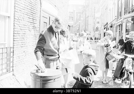 Boy Scout Spendenaktion Haarlem the Netherlands, Whizgle News, Dutch Desk, Niederlande, 1950 - 2000 auf 05-1967. Diese Themen sind in der Abbildung dargestellt. Die Szene zeigt eine lebhafte Straße voller Aktivität an einem hellen Tag. Das Sonnenlicht fließt auf den Pflastersteinpflaster, wirft weiche Schatten und verbessert die Lebendigkeit der Umgebung. Im Vordergrund beugt sich ein Mann in einer stilvoll geschnittenen Pullover-Weste über einen Mülleimer und kümmert sich aktiv um die Entsorgung von Gegenständen. Seine Brille reflektiert einen Lichtblick, was auf Fokus und Absicht hindeutet. In der Nähe steht ein Junge im dunklen Mantel nahe Stockfoto