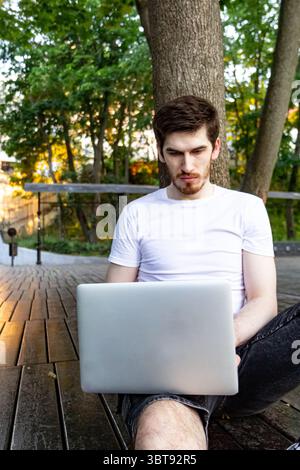 Ein lässiger Mann sitzt auf Holzdeck, Rücken an den Baum und tippt am Laptop. Frontwinkel, friedliche Parkatmosphäre, mit weißem Hemd und dunklen Shorts. Stockfoto