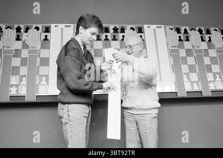 H.S.T. : Grandmaster B Draw, Whizgle News, Dutch Desk, Niederlande, 1950 - 2000 am 12.01.1992. Dies sind die Elemente im Bild. Die Szene zeigt zwei Personen, die sich in einer lebhaften Interaktion engagieren. Eine Person, ein junger Mann, steht links. Er hat eine helle, leicht zerzauste Frisur und trägt einen gemütlichen, strukturierten Pullover mit auffälligem Muster, kombiniert mit Jeans. Sein Ausdruck vermittelt eine Mischung aus Enthusiasmus und Konzentration, da er einen langen Streifen Papier hält, auf dem Zahlen sichtbar sind. Auf der rechten Seite lächelt ein älterer Herr weit, strahlt Wärme und ein Gefühl von p aus Stockfoto