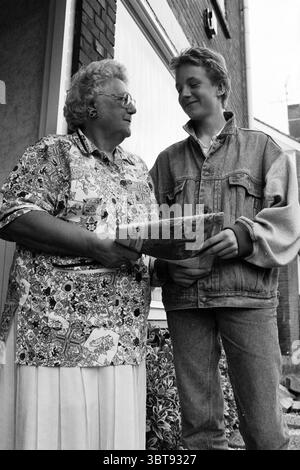 Frau Jansen + Zeitungsberichterstatter Nieuw-Vennep Nieuw-Vennep, Whizgle News, Dutch Desk, Niederlande, 1950 - 2000 am 15-09-1993. Das Bild enthält diese Themen. In einer bezaubernden Umgebung im Freien stehen zwei Personen im Gespräch. Die ältere Frau strahlt Wärme aus mit ihren lockigen, silbernen Haaren und einem breiten Lächeln, gekleidet in einer hell gemusterten Bluse, die einen Kontrast zu den gedämpften Tönen ihrer Umgebung bildet. Ihr Outfit besticht durch eine Mischung aus floralen und geometrischen Designs, die leuchtende Farben wie Pink, Blau und Gelb zeigen, die eine lebendige Persönlichkeit vermitteln. Neben ihr strahlt ein junger Mann aus Stockfoto