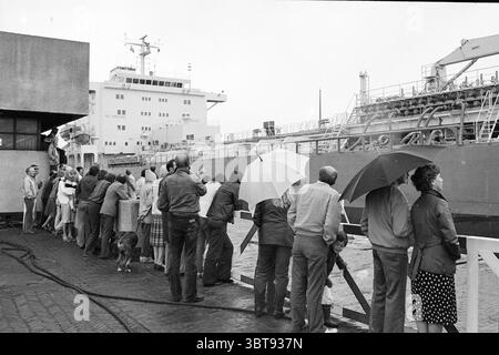 Schlechtes Wetter IJmuiden Wetter Wetter Service IJmuiden Velsen IJmuiden Niederlande, Whizgle News, Dutch Desk, Niederlande, 1950 - 2000 am 21-07-1981. Das Bild enthält diese Themen. Eine Gruppe von Einzelpersonen steht neben einem Dock und blickt alle aufmerksam auf ein großes Schiff, das in der Nähe vertäut ist. Die Gruppe, die sich aus Männern und Frauen zusammensetzt, zeigt eine Reihe von Ausdrucksformen, von Neugier bis hin zur Begeisterung. Verschiedene Regenschirme, einige geschlossen und andere offen, bieten einen Spritzer weißer und heller Töne vor dem Hintergrund grauer Himmel, was auf einen drohenden oder anhaltenden Nieselregen hindeutet. Die Leute sind in einer Mixtur gekleidet Stockfoto
