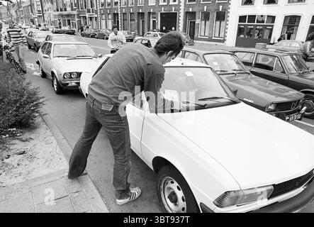 Promotion ENFB Radwege Radfahrer, Whizgle News, Dutch Desk, Niederlande, 1950 - 2000 am 20-08-1980. Die Abbildung zeigt diese Themen. In der Szene konzentriert sich ein junger Mann auf ein weißes Auto, das entlang einer engen Straße geparkt ist. Er beugt sich leicht über das Fahrzeug und überprüft den Bereich zwischen Windschutzscheibe und Motorhaube. Sein dunkler Strickpullover bildet einen Kontrast zum hellen Finish des Autos, während seine Jeans einen lässigen Look vervollständigen. Die Umgebung bietet eine Sammlung von Oldtimern, die auf beiden Seiten der Straße in einer Reihe von Farben, darunter gedämpfte c-Töne, angeordnet sind Stockfoto