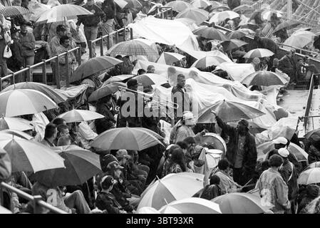 HHW Rain [Haarlem Baseball Week], Whizgle News, Dutch Desk, Niederlande, 1950 - 2000 am 28-06-1996. Dies sind die Elemente im Bild. Die Szene zeigt eine überfüllte Umgebung im Freien, wahrscheinlich ein Stadion, gefüllt mit einer vielfältigen Gruppe von Menschen, die sich unter einem trüben, bedeckten Himmel versammelt haben. Zahlreiche Regenschirme schaffen einen lebendigen Farbteppich, mit hellen Rot-, Blau-, Gelb- und Grüntönen, die sich von der gedämpften grauen Umgebung abheben. Einige Regenschirme sind von innen nach außen geklappt oder haben ein paar Regentropfen, die auf schlechtes Wetter hinweisen. Die Teilnehmer sind in einer Mischung aus Casua gekleidet Stockfoto