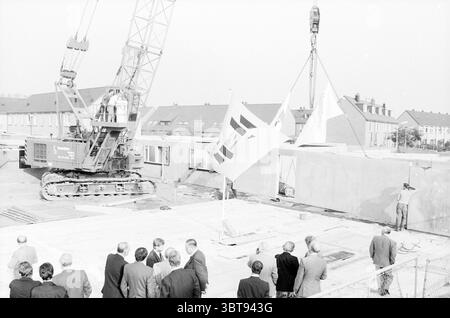 First Pile Weversland Noordwijkerhout First Pile Noordwijkerhout, Whizgle News, Dutch Desk, Niederlande, 1950 - 2000 am 18-10-1989. Die Abbildung zeigt diese Themen. In einer belebten Bauszene ragt ein großer Kran über einer Gruppe elegant gekleideter Individuen, die sich auf einer Ebenen Fläche versammeln, die wie die Grundlage eines Gebäudes zu sein scheint. Der Kran mit einem langen Ausleger, der sich nach oben erstreckt, hebt gerade große Betonplatten an, die in der Luft prekär ruhen. Die Farben der Maschinen stehen in starkem Kontrast zu den gedämpften Umgebungstönen; der Kran ist besonders industriell Stockfoto