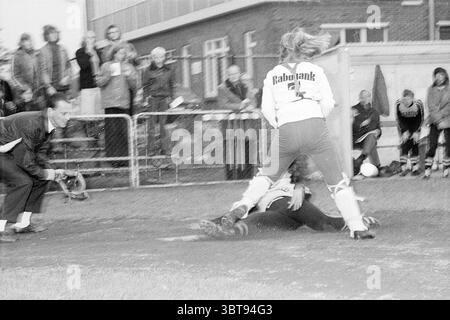Terrasvogels - Seleritas Softball, Whizgle News, Dutch Desk, Niederlande, 1950 - 2000 am 27-04-1974. Das Bild enthält diese Themen. In der Szene spielen zwei Spieler während eines Fußballspiels einen dynamischen Moment. Ein Spieler, der in einem weißen Oberteil mit einem auffälligen Logo gekleidet ist, befindet sich in einer gehockerten Position und versucht, auf dem Ball zu spielen. Der andere Athlet, der ein dunkleres Trikot trägt, ist am Boden und versucht, der Herausforderung zu entgehen oder sie zu bekämpfen. Der Boden besteht aus einer Mischung aus Gras und Schmutz, was auf starke Beanspruchung und möglicherweise Regen in jüngster Zeit hinweist, was der Umgebung eine leicht schlammige Textur verleiht. Sur Stockfoto