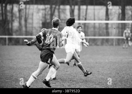Football Match, Whizgle News, Dutch Desk, Niederlande, 1950 - 2000 am 12-1988. Diese Themen werden in der Abbildung angezeigt. Die Szene fängt einen dynamischen Moment auf einem Fußballfeld ein, der durch eine Mischung aus Bewegung und Fokus gekennzeichnet ist. Im Mittelpunkt der Komposition stehen zwei Spieler in der Mitte der Action. Der eine trägt ein leuchtendes rotes Trikot mit der Nummer 10 und zeigt entschlossene Leichtathletik, während er über das üppige grüne Gras sprintet. Sein Haar ist leicht zerzaust, was auf die Intensität des Spiels hindeutet. Der gegnerische Spieler, der in einem weißen Trikot gekleidet ist, wird weiter positioniert, wobei die Nummer 5 auf dem Rücken sichtbar ist. Sein Stockfoto