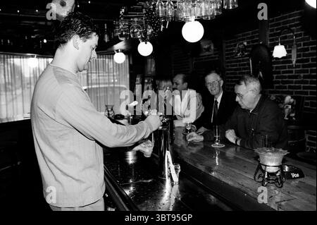 Café de Tapperij, Whizgle News, Dutch Desk, Niederlande, 1950 - 2000 am 27-02-2001. Das Bild enthält diese Themen. In einer gemütlichen, schwach beleuchteten Bar ist die Atmosphäre warm und einladend, die durch satte Holztöne und das sanfte Leuchten der Hängeleuchten vermittelt wird. Ein junger Barkeeper in legeren Kleidern konzentriert sich darauf, ein Getränk aus einem glänzenden Wasserhahn zu gießen. Seine Haltung deutet auf Hingabe an sein Handwerk hin, mit einer leichten Neigung zur Theke, die seine Auseinandersetzung mit der anstehenden Aufgabe unterstreicht. Die Bar selbst ist poliert und reflektierend, mit einer Reihe sauberer Gläser, die ordentlich über der Theke gestapelt sind. Be Stockfoto