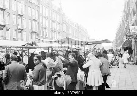 Bericht Albert Kuipstraat Report, Whizgle News, Dutch Desk, Niederlande, 1950 - 2000 am 22-05-1973. Die Abbildung zeigt diese Themen. Die Szene fängt einen belebten Straßenmarkt mit Verkaufsständen ein und schafft eine lebhafte Atmosphäre voller Aktivität. Auf beiden Seiten ragen Reihen von hohen, schmalen Gebäuden mit klassischen architektonischen Merkmalen über die Szene, deren Fassaden durch eine Mischung gedämpfter Töne gekennzeichnet sind. Auf dem Markt leben Menschen unterschiedlicher Stile und Alters, die sich mit Einkaufen und Sozialen beschäftigen; einige untersuchen Waren, während andere lebhaft miteinander reden. Helle, gemusterte Stoffe A Stockfoto