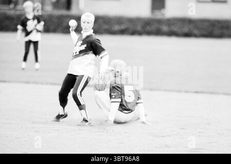 HHC - Heiloo Softball, Whizgle News, Dutch Desk, Niederlande, 1950 - 2000 am 12-08-1987. Dies sind die Elemente im Bild. Die Szene fängt einen Moment auf einem Baseballfeld während eines Spiels ein. Im Vordergrund steht ein Spieler in einem hellen Trikot mitten im Wurf und zeigt eine athletische Haltung. Ihr Ausdruck ist bestimmt, und sie haben kurze, helle Haare. Dieser Spieler trägt schwarze Leggings, die mit weißen Streifen verziert sind und für ein dynamisches Flair sorgen. Auf dem Boden sitzt ein weiterer Spieler, der in einem hellroten Trikot gekleidet ist, wobei die Nummer 5 deutlich sichtbar dargestellt wird. Sie sind in einer defensiven Position, Stockfoto