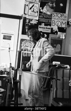 Konzert Hannes Kuiper Concerts Music, Whizgle News, Dutch Desk, Niederlande, 1950 - 2000 am 19-11-1983. Diese Themen werden in der Abbildung angezeigt. Die Szene zeigt einen schwach beleuchteten Raum mit nostalgischem Flair. Die Kulisse ist mit mehreren Plakaten mit farbenfrohen künstlerischen Designs und auffälliger Typografie geschmückt, die auf eine kreative oder musikalische Atmosphäre hinweisen. Die Wände scheinen etwas abgenutzt zu sein, was das Gesamtgefühl von Alter und Charakter noch verstärkt. Im Vordergrund steht eine Person mit langen, dunklen Haaren nachdenklich. Sie tragen eine lässige helle Jacke über einem dunkleren Hemd, kombiniert mit einer helleren Hose, cr Stockfoto