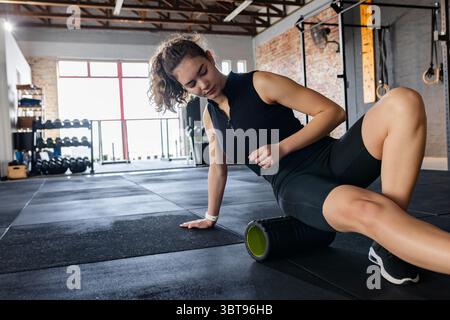 Erwachsene weibliche Fitness-Enthusiasten sitzen auf einer Turnmatte mit Schaumstoffrolle am Oberschenkel in der Nähe des Hantelhalters Stockfoto
