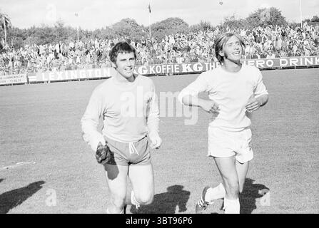 Telstar Football Match, Whizgle News, Dutch Desk, Niederlande, 1950 - 2000 am 09-1970. Dies sind die Themen im Bild. In einem belebten Outdoor-Stadion joggen zwei männliche Athleten auf einem grasbewachsenen Feld. Die Kulisse zeigt eine große Zuschauermenge, deren Jubel in der Luft nachhallt. Der Himmel ist eine Mischung aus weichen, flauschigen Wolken und blauen Flecken, die auf einen angenehmen Tag hindeuten. Der erste Athlet, der in Shorts und einem leichten Oberteil gekleidet ist, strahlt Selbstvertrauen aus. Sein kurzes Haar ist leicht zerzaust, und ein breites Lächeln zeigt seine Zufriedenheit. Neben ihm, der zweite Athlet, sp Stockfoto