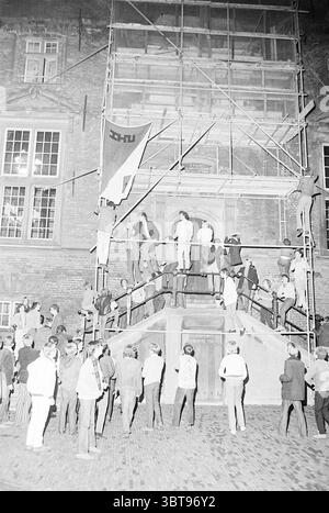 Riots Grote Markt Grote Markt Riots Haarlem Grote Markt The Netherlands, Whizgle News, Dutch Desk, Niederlande, 1950 - 2000 am 12-09-1967. Die Abbildung zeigt diese Themen. In einer geschäftigen Szene in der Nacht in der Nähe eines imposanten Gebäudes versammelt sich eine Menschenmenge um ein Gerüst, das an der Fassade errichtet wurde. Die Architektur hinter dem Gerüst zeichnet sich durch große, bogenförmige Fenster aus, die komplizierte Details zeigen, die auf seine historische Bedeutung hinweisen. Die Menge besteht aus einer vielfältigen Mischung von Individuen, einige klettern auf das Gerüst, andere stehen auf den Stufen unten, Stockfoto