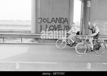 Defacing viaduct Hoofddorp Texte Hoofddorp the Netherlands, Whizgle News, Dutch Desk, Niederlande, 1950 - 2000 am 08-06-1989. Das Bild enthält diese Themen. In der Szene fahren zwei Personen auf einer Straße, die unter einer großen Betonüberführung verläuft. Die Atmosphäre hat ein Gefühl von ungezwungener Freizeit, da die Radfahrer ihre Fahrt zu genießen scheinen, wobei eine Person spielerisch mit erhobener Hand Gesten gibt. Die Straße besteht aus grauem Asphalt, und der Hintergrund zeigt eine weitläufige, offene Landschaft mit subtilen, sanften Hügeln in sanften Grün- und Brauntönen, was auf eine ruhige Grabung hindeutet Stockfoto