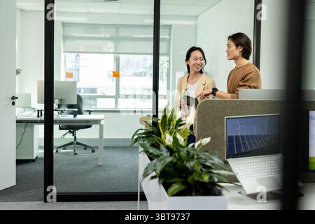 Asiatische Kollegen halten ein Tablet in der Hand, während sie über erneuerbare Energien auf einem Laptop neben Pflanzmaschine am Schreibtisch sprechen Stockfoto