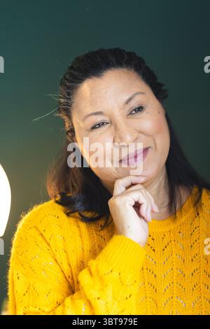 Frau mit gelbem Pullover, das Kinn auf der Hand liegend, während sie außerhalb der Kamera im privaten Zimmer mit Lampe blickte Stockfoto