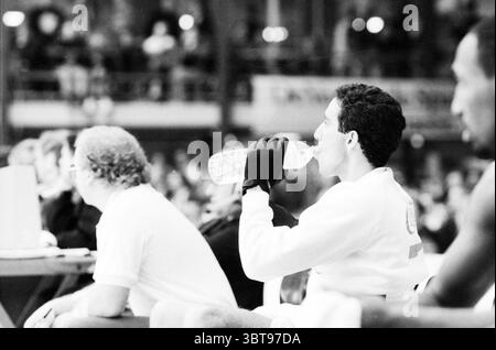 Levi's-Commodore Basketballspiel, Whizgle News, Dutch Desk, Niederlande, 1950 - 2000 am 27.12.1989. Dies sind die Themen im Bild. In einer dynamischen Sportumgebung werden mehrere Personen in einem Moment der Vorfreude erfasst. Die Komposition zeigt einen jungen Mann im Vordergrund, der einen Schluck aus einer durchsichtigen Plastikwasserflasche nimmt. Er trägt ein leichtes, langärmeliges Sportoberteil und schwarze Handschuhe, die einen starken Kontrast zu den umgebenden Elementen bilden. Sein Ausdruck ist fokussiert und deutet sowohl auf Konzentration als auch auf ein Bedürfnis nach Hydration während einer Pause hin. Der Hintergrund zeigt verschwommene Outl Stockfoto