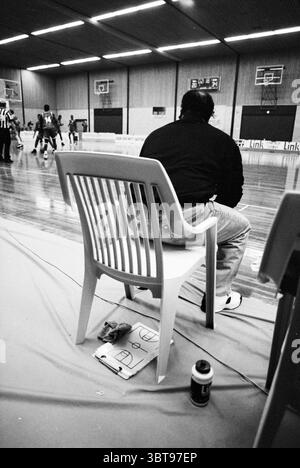 Akrides - Goba Basketball, Whizgle News, Dutch Desk, Niederlande, 1950 - 2000 am 06-01-1995. Diese Themen werden in der Abbildung angezeigt. In der Szene erwacht ein geräumiges Indoor-Gymnasium mit Aktivität zum Leben. Der Hartholzboden ist poliert und glänzt unter den hellen Deckenleuchten, die einen warmen Farbton reflektieren, der sich mit den kühlen Tönen der Umgebung abhebt. Die Holzwände schaffen eine gemütliche, aber lebendige Atmosphäre, die durch das leise Echo von quietschenden Turnschuhen und das Geräusch eines hüpfenden Basketballs verziert wird. Im Vordergrund befindet sich eine Einzelfigur auf einem weißen Plastikstuhl, der leicht außermittig positioniert ist. Stockfoto