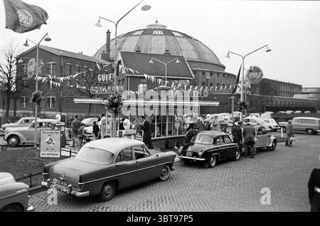 Eröffnung Schmier-/Tankstelle GULF Brinkmann Haarlem Opening Opening Opening Opening Commissioning, Whizgle News, Dutch Desk, Niederlande, 1950 - 2000 am 26-11-1960. Dies sind die Themen im Bild. Die Szene spielt in einer lebhaften städtischen Umgebung aus einer vergangenen Zeit, wahrscheinlich Mitte des 20. Jahrhunderts. Der Schwerpunkt ist ein beeindruckendes, großes, gewölbtes Gebäude im Hintergrund mit einem unverwechselbaren Dach und einem architektonischen Stil, der seine Pracht unterstreicht. Im Vordergrund säumen mehrere Oldtimer eine kopfsteingepflasterte Straße, die eine Reihe älterer Modelle mit abgerundeten Kanten und Chromdetai zeigen Stockfoto