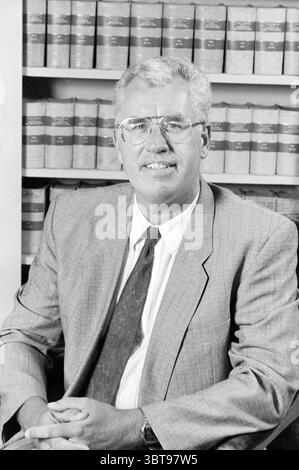 Porträt eines Mannes (aus Anwaltskanzlei) Haarlem Grote Markt 2 Niederlande, Whizgle News, Dutch Desk, Niederlande, 1950 - 2000 im Jahr 1990. Die Abbildung zeigt diese Themen. In einer professionellen Umgebung sitzt ein Mann selbstbewusst und strahlt einen Hauch von Autorität aus. Er hat helle Haare, ordentlich gekämmt und trägt einen gut geschnittenen Anzug in Hellgrau mit dezenten Mustern, ergänzt durch ein knuspriges weißes Hemd und eine gemusterte Krawatte in warmen Tönen. Seine runde Brille liegt auf seiner Nase und bildet einen freundlichen und zugänglichen Ausdruck. Der Hintergrund zeigt ordentlich angeordnete Gesetzbücher, deren Wirbelsäule einen Verlauf von zeigt Stockfoto