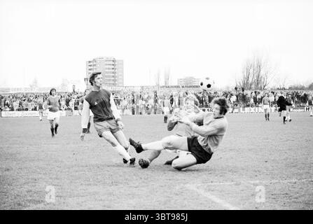 Haarlem - SVV Football Haarlem, Whizgle News, Dutch Desk, Niederlande, 1950 - 2000 am 23-04-1972. Die Abbildung zeigt diese Themen. Die Szene fängt einen intensiven Moment auf einem grasbewachsenen Fußballfeld mit voller Aktivität ein. Der Hauptfokus liegt auf zwei Spielern in der Nähe der Mitte des Spielfeldes, die in einem dynamischen Spiel spielen. Der eine trägt ein knallrotes Trikot und ist bereit, den Ball zu kicken, während der andere, der eine graue Torwartuniform trägt, auf dem Boden liegt, um ihn abzufangen. Ihre Körper sind in Aktion positioniert und vermitteln sowohl Anstrengung als auch Spannung. Um sie herum gibt es zusätzliche Spieler, mit Stockfoto