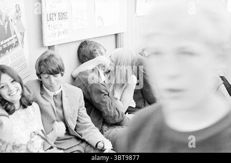 Bericht Stuif Stuif Stuif, Whizgle News, Dutch Desk, Niederlande, 1950 - 2000 am 04-07-1967. Die Abbildung zeigt diese Themen. In einem lebendigen sozialen Umfeld versammeln sich mehrere Personen und schaffen eine Atmosphäre jugendlicher Energie und Intimität. Auf der einen Seite lächelt eine junge Frau mit langen, dunklen Haaren und einem gemusterten Kleid breit, ihr Ausdruck strahlt Freude aus. Neben ihr sitzt ein junger Mann, der einen Blazer und ein Hemd mit Kragen trägt, der zurückhaltender erscheint und ein nachdenkliches Verhalten aufweist. In der Mitte teilen sich zwei Personen einen kurzen Moment, gefangen in einer Umarmung. Die Frau, gekennzeichnet von langem Blo Stockfoto