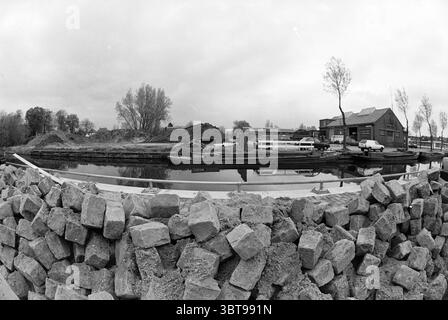 Renovierung des Hafens in Hillegom Hillegom, Whizgle News, Dutch Desk, Niederlande, 1950 - 2000 am 27-04-1992. Diese Themen sind in der Abbildung dargestellt. Die Szene spielt an einem ruhigen Gewässer, der den bewölkten Himmel darüber reflektiert, der in sanften Grau- und Weißtönen gemalt ist. Auf der rechten Seite ist eine Ansammlung bescheidener Gebäude mit einfachen Dächern zu sehen, deren Oberflächen verwittert und gedämpft sind und sich leicht in die graue Atmosphäre einfügen. Diese Strukturen scheinen zweckmäßig zu sein, möglicherweise Lagerhäuser oder Lagereinrichtungen, die mit dem nahegelegenen Wasserweg in Verbindung stehen. Im Vordergrund befindet sich eine niedrige Steinbarriere aus r Stockfoto