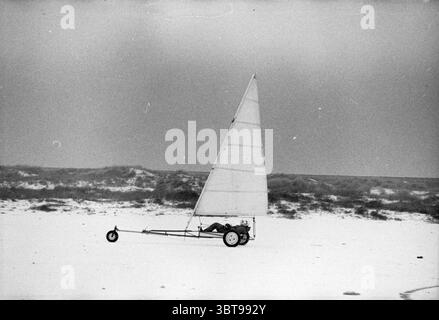 Atmosphäre Strand IJmuiden mit Schnee Schnee IJmuiden Niederlande, Whizgle News, Dutch Desk, Niederlande, 1950 - 2000 am 01-01-1986. Dies sind die Themen im Bild. Die Szene zeigt eine einsame Landyacht, die über einen ausgedehnten Strandabschnitt gleitet, der sich durch seinen glatten, weißen Sand auszeichnet. Die Landyacht verfügt über ein großes dreieckiges Segel, das straff und gegen eine sanfte Brise geneigt ist und einen auffälligen Kontrast zum gedämpften grauen Himmel bietet. Sein einfacher Rahmen wird von zwei Rädern gestützt, sodass er leicht manövrieren kann. Im Hintergrund sind sanfte, hügelige Dünen sichtbar, ihre Kurven Stockfoto